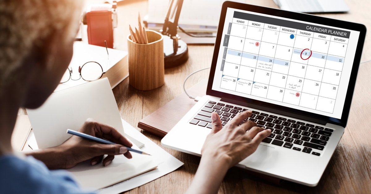 Closeup of desk and a woman’s hand on a laptop keyboard as she looks at a schedule planner on the computer.